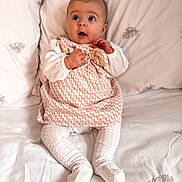 Lainey joined the competition — help win amazing prizes! baby, infant, child, dress, white_clothing, patterned_fabric, bed, pillow, leaf_pattern, soft_texture, sitting, curious_expression, wide_eyes, hands, legs, indoors, portrait, cute, cozy, comfortable