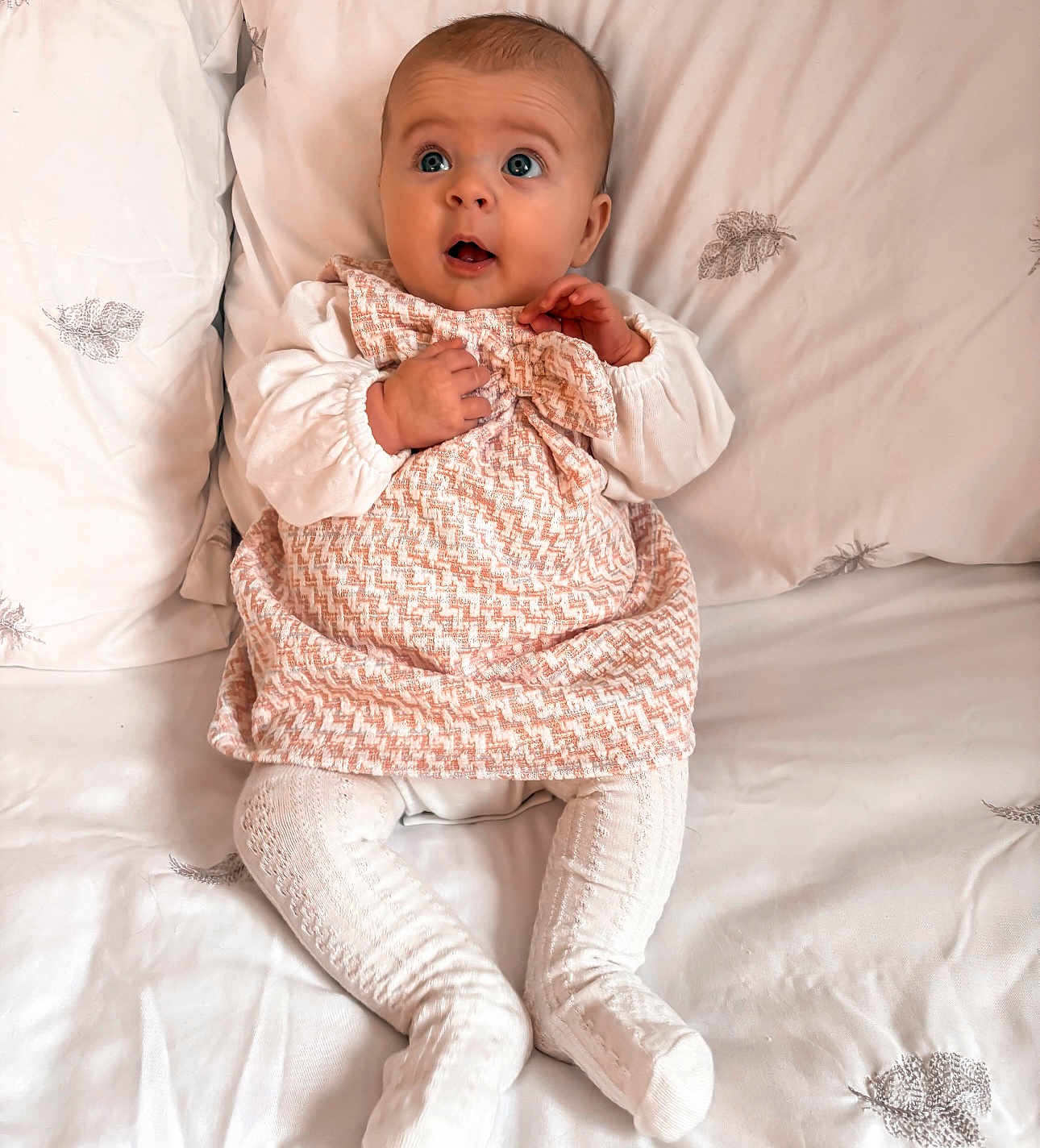 Lainey joined the competition — help win amazing prizes! baby, infant, child, dress, white_clothing, patterned_fabric, bed, pillow, leaf_pattern, soft_texture, sitting, curious_expression, wide_eyes, hands, legs, indoors, portrait, cute, cozy, comfortable