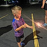 Gunnar is registered to the contest to win money with this photo: child, sparkler, night, parking_lot, street_light, asphalt, shorts, tshirt, sandals, adult, hand, sparkle, shadow, tree, light, expression, curious, outdoor, person, fun
