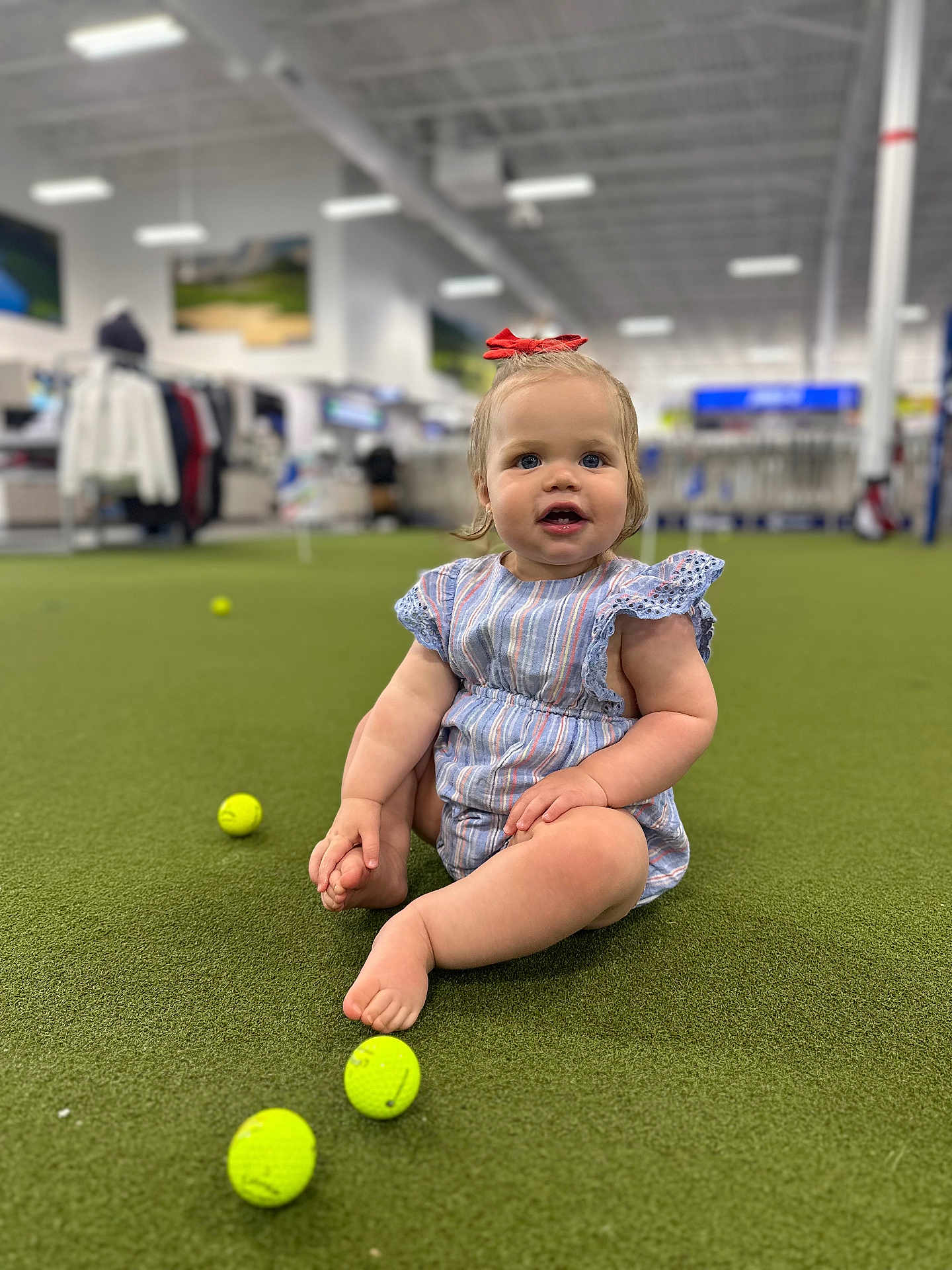 Navy joined the competition — help win amazing prizes! toddler, child, indoor, green_turf, golf_balls, barefoot, red_bow, blue_eyes, striped_clothing, sitting, curious_expression, short_hair, playing, sports_store, blurred_background, person, floor, cute, casual, young_child