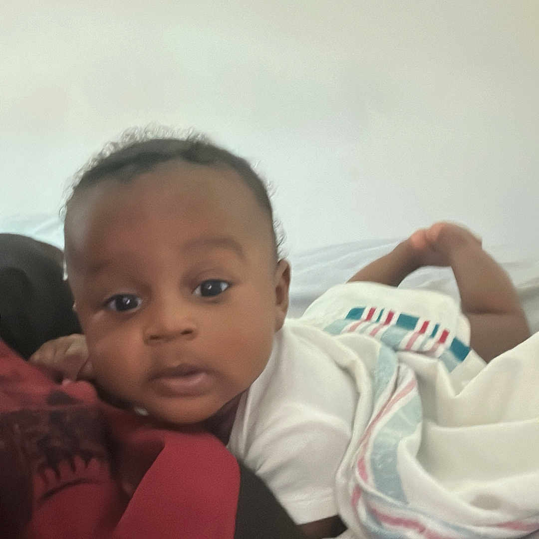 Masiyah is registered to the contest to win money with this photo: baby, baby_blanket, bed, blanket, child, close_up, cozy, curious, dark_skin, face, head, indoor, infant, looking_at_camera, lying_down, portrait, resting, short_hair, soft_lighting, white_clothing