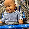 toddler, child, shopping_cart, grocery_store, smiling, blue_clothing, aisle, floor, hand, face, person, portrait, indoor, happy, retail, baby, cart_handle, store_shelf, shopping, casual