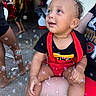 baby, background, barefoot, black_shirt, bubbles, casual, child, curly_hair, daylight, earrings, face, hand, lap, legs, outdoor, people, person, red_clothing, smiling, toddler