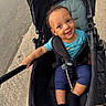 Kairo is registered to the contest to win money with this photo: baby, black_socks, blue_shirt, casual_clothing, child, curly_hair, cute, happy, holding, navy_pants, outdoor, pavement, person, playful, seatbelt, smile, stroller, sunlight, toddler, walking