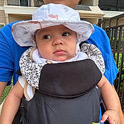 Zakkai is registered to the contest to win money with this photo: baby, child, hat, carrier, person, blue_shirt, outdoor, fence, greenery, curious, face, headwear, infant, holding, sunhat, patterned, closeup, daylight, person_holding_baby, casual