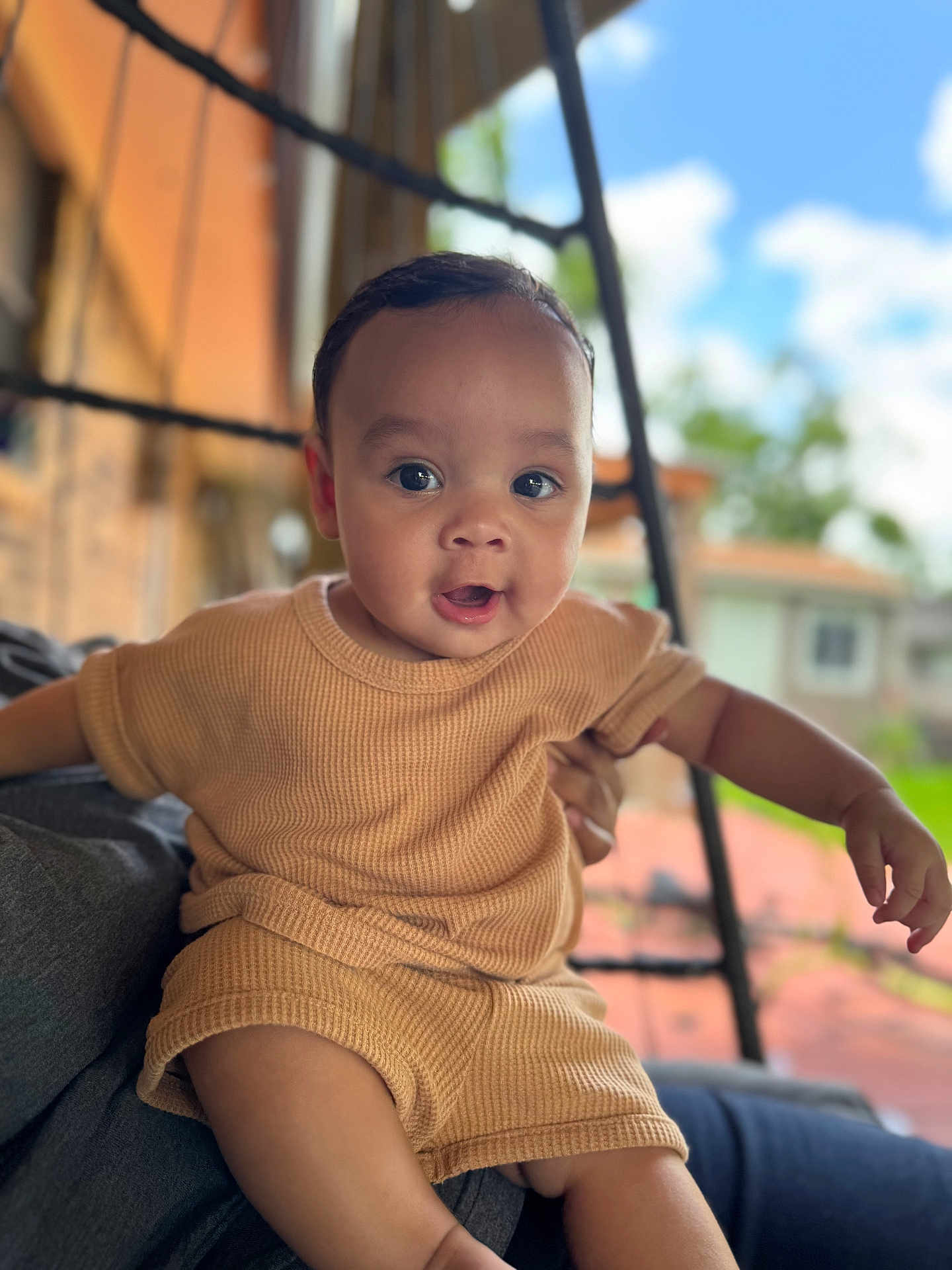 Zakkai is registered to the contest to win money with this photo: baby, child, infant, person, outdoor, clothing, tan_outfit, face, eyes, mouth, skin, hand, sitting, blurred_background, sky, greenery, daylight, portrait, cute, expression