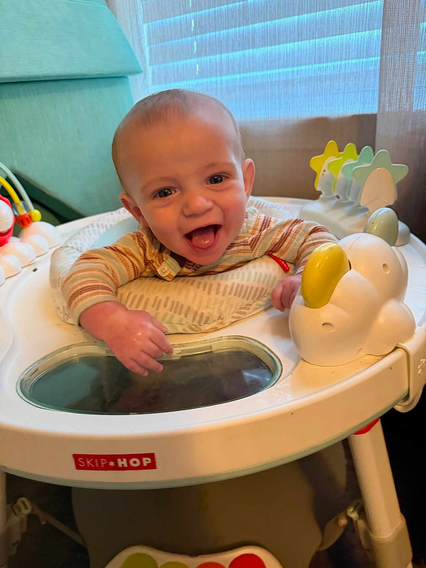 Jayden is registered to the contest to win money with this photo: baby, child, smiling, laughing, activity_center, toy, indoor, playtime, infant, happy, cute, person, seat, colorful, furniture, curtains, clothing, hands, face, expression