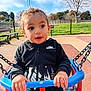 child, toddler, swing, playground, outdoor, park, fence, bench, grass, sky, clouds, person, jacket, nike, hands, face, expression, daylight, casual_clothing, chains