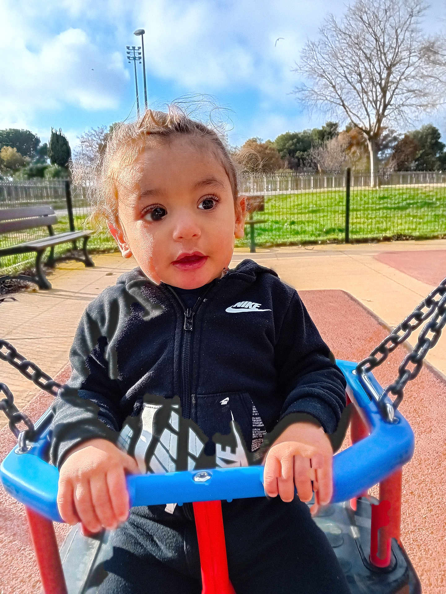 Kais participe au concours pour gagner de l'argent avec cette photo : child, toddler, swing, playground, outdoor, park, fence, bench, grass, sky, clouds, person, jacket, nike, hands, face, expression, daylight, casual_clothing, chains