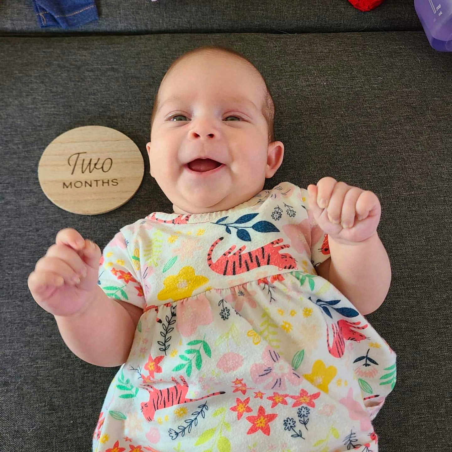 Oriana joined the competition — help win amazing prizes! baby, celebration, child, clothing, colorful, cute, dress, fabric, fist, floral_pattern, happy, indoors, infant, lying_down, person, portrait, smiling, tiger_print, two_months, wooden_plaque