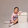 child, girl, sitting, pink_sweatshirt, plaid_skirt, white_socks, white_shoes, ottoman, neutral_background, floor, smile, casual_clothing, indoor, portrait, young_child, fashion, relaxed_pose, studio_lighting, short_hair, cute