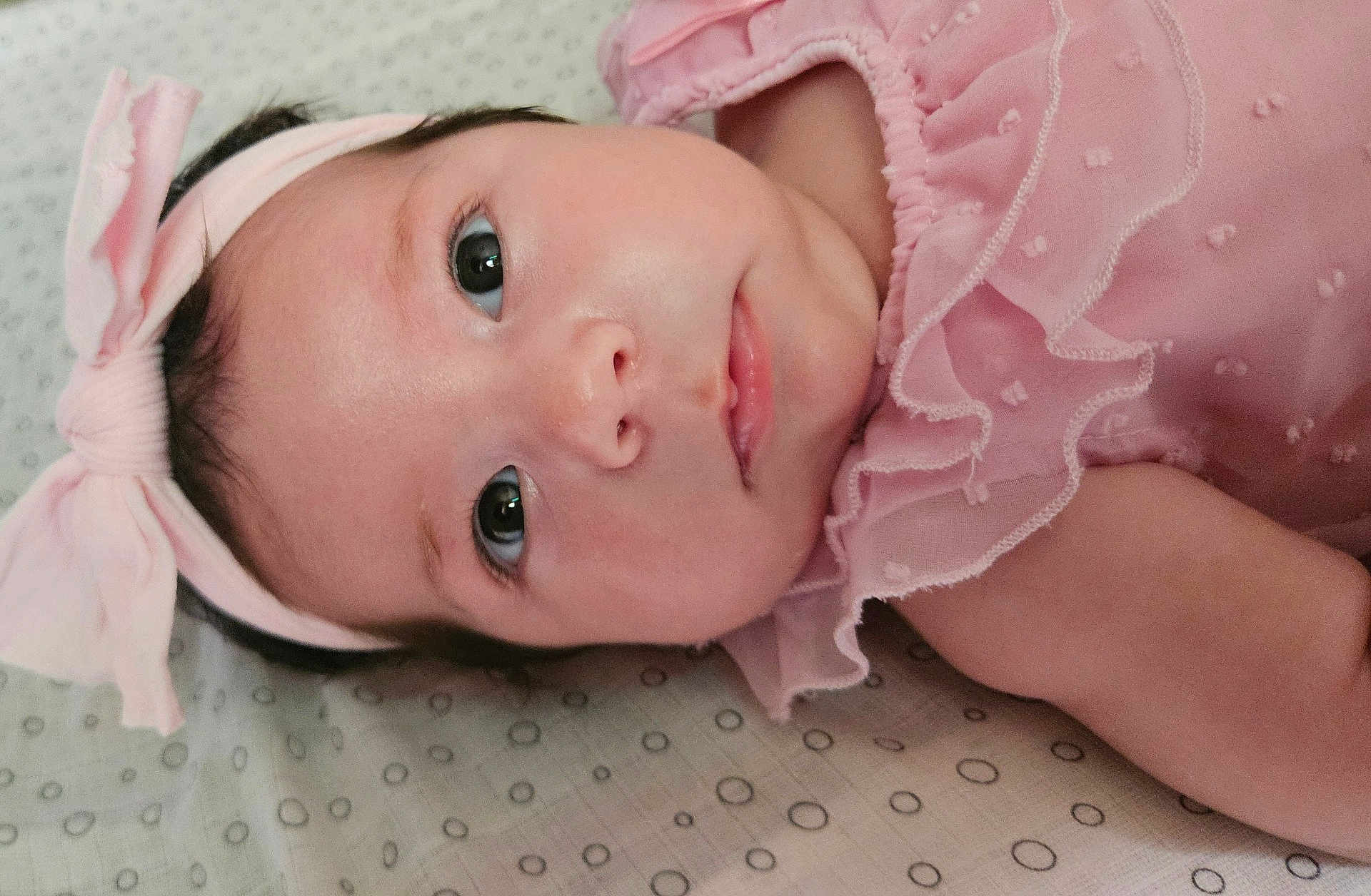 Aury is registered to the contest to win money with this photo: baby, infant, face, headband, pink_clothing, bow, lying_down, close_up, child, cute, skin, eyes, portrait, young_child, bed, fabric, pattern, soft, person, adorable