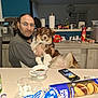man, dog, table, coffee_cup, smartphone, glass, snacks, kitchen, paper_towels, glasses, face, furniture, indoor, pet, holding, seated, appliance, counter, casual, relaxed