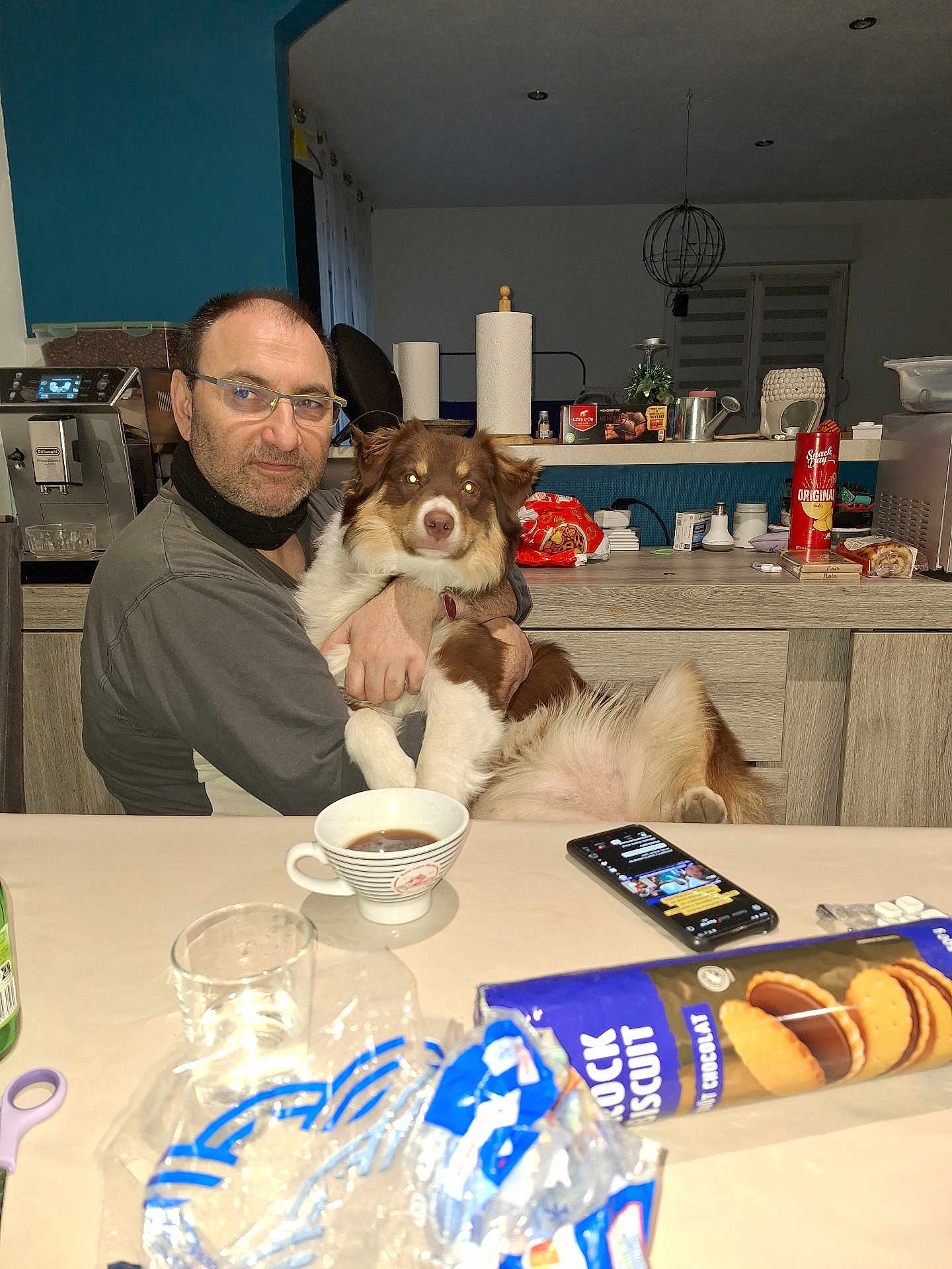 Enya a rejoint le concours — aidez-le/la à gagner de superbes lots ! man, dog, table, coffee_cup, smartphone, glass, snacks, kitchen, paper_towels, glasses, face, furniture, indoor, pet, holding, seated, appliance, counter, casual, relaxed