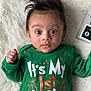 baby, blanket, calendar_block, celebration, child, clothing, cute, expression, face, first_birthday, green_shirt, hair, hand, indoor, infant, lying_down, new_year, portrait, text_on_clothing, wide_eyes