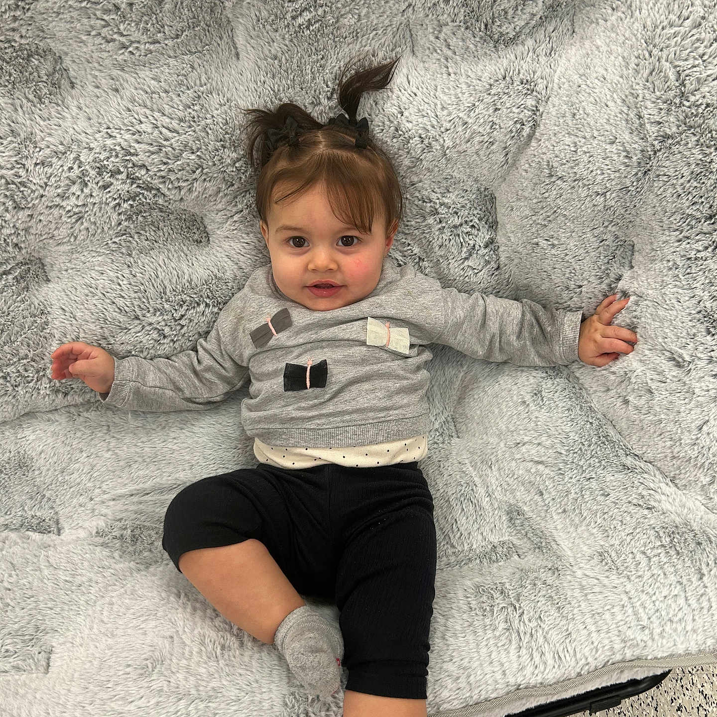Alaia is registered to the contest to win money with this photo: baby, black_pants, blanket, child, clothing, cute, face, floor, girl, gray_sweater, hair, indoor, person, pigtails, portrait, relaxed, smiling, socks, soft_texture, toddler