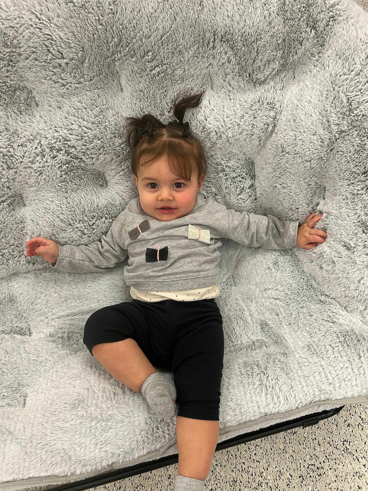 Alaia is registered to the contest to win money with this photo: toddler, child, baby, girl, smiling, face, hair, pigtails, gray_sweater, black_pants, socks, blanket, soft_texture, indoor, floor, cute, relaxed, portrait, clothing, person