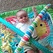 Emmett is registered to the contest to win money with this photo: baby, play_mat, toy, infant, carpet, colorful, striped, clothing, person, face, legs, footwear, play_gym, cute, indoors, child, smile, milk_mustache, soft_toys, relaxing