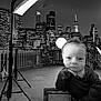 baby, infant, face, cityscape, night, black_and_white, studio_light, urban, railing, photography, portrait, expression, hand, clothing, building, skyline, light_stand, outdoor, monochrome, person
