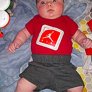Rowan is registered to the contest to win money with this photo: baby, infant, red_shirt, gray_shorts, white_socks, play_mat, toys, blanket, patterned_background, indoor, child, person, cute, lying_down, wide_eyes, chubby_arms, soft_lighting, expression, close_up, home