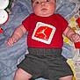 baby, infant, red_shirt, gray_shorts, white_socks, play_mat, toys, blanket, patterned_background, indoor, child, person, cute, lying_down, wide_eyes, chubby_arms, soft_lighting, expression, close_up, home