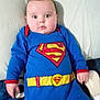 baby, blanket, blue_onesie, bottle, child, chubby_cheeks, clothing, couch, cute, face, hand, human, indoor, infant, lying_down, pillow, portrait, relaxed, superman_costume, young_child