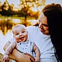 baby, woman, smiling, outdoor, nature, water, trees, sunset, golden_hour, happy, holding, person, infant, casual_clothing, denim, portrait, family, love, warm_light, blurred_background