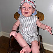 Kashton joined the competition — help win amazing prizes! baby, child, smiling, hat, striped_clothing, romper, wooden_chair, indoor, happy, feet, hands, face, person, cute, infant, sitting, floor, wood_floor, bracelet, red_eyes