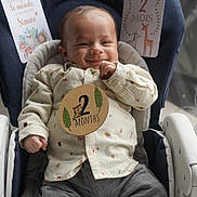 Simão a rejoint le concours — aidez-le/la à gagner de superbes lots ! baby, infant, smile, clothing, shirt, pants, baby_seat, milestone_card, text, hand, face, cute, child, indoor, comfort, happy, young, portrait, sitting, cozy