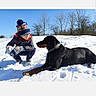 Orko a rejoint le concours — aidez-le/la à gagner de superbes lots ! child, dog, snow, winter_clothing, hat, gloves, outdoor, blue_sky, trees, sunlight, playful, pet, crouching, lying_down, nature, cold_weather, friendship, smiling, animal, daytime