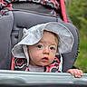 baby, child, infant, stroller, sun_hat, outdoor, portrait, face, eyes, wide_eyed, sitting, hand, gripping, floral_clothing, patterned_clothing, greenery, grass, daylight, closeup, adorable