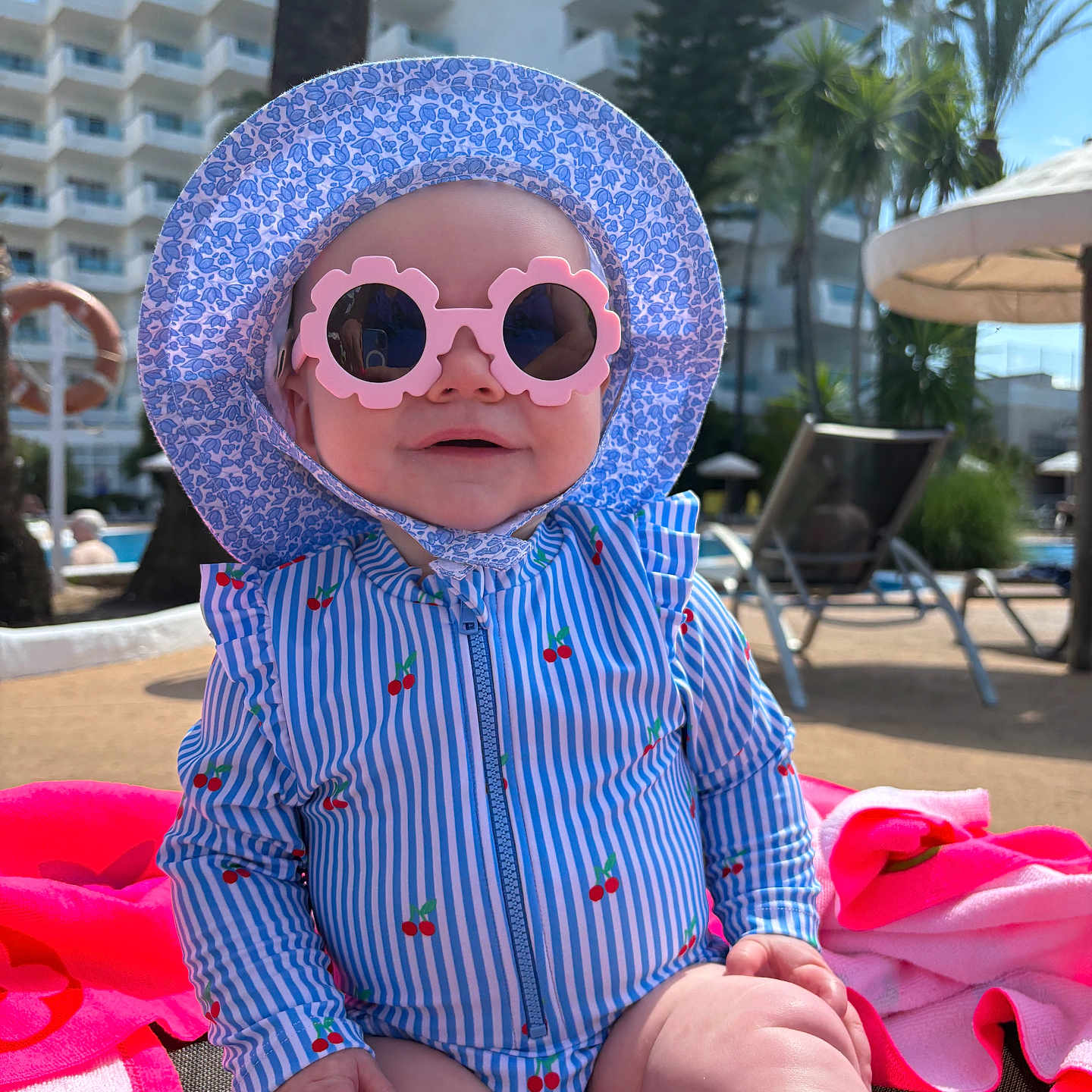 Arwen is registered to the contest to win money with this photo: baby, cherry_pattern, child, hat, holiday, lounge_chair, outdoor, person, poolside, relaxation, smiling, striped_clothing, summer, sunglasses, sunhat, sunny, swimsuit, toddler, trees, vacation