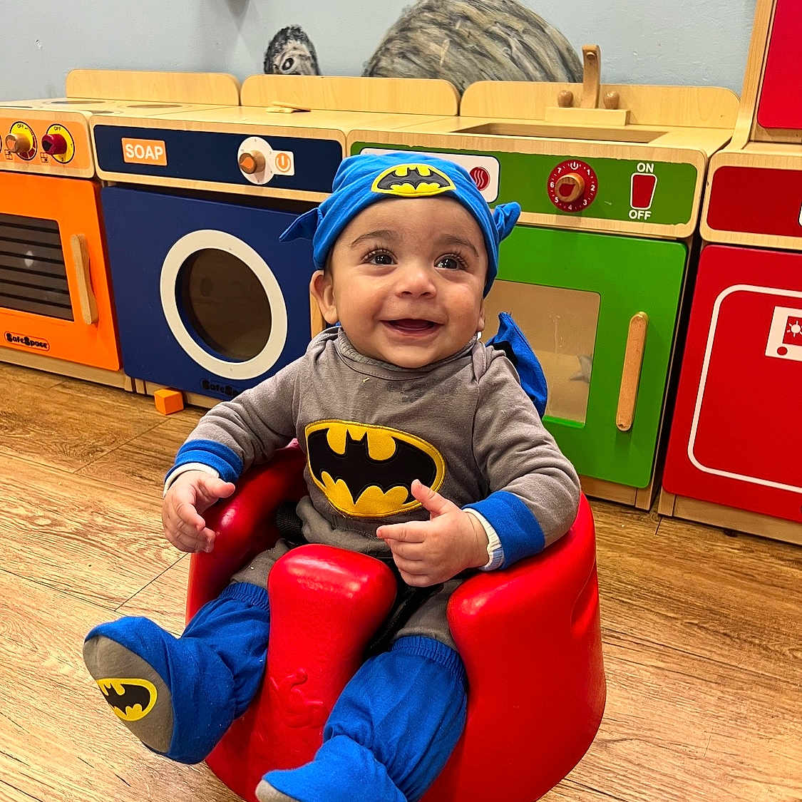 Danny is registered to the contest to win money with this photo: baby, batman_costume, child, colorful, costume, cute, footwear, happy, headwear, indoors, infant, person, play, playroom, red_baby_seat, seat, smiling, toy_kitchen, wooden_floor, young_child