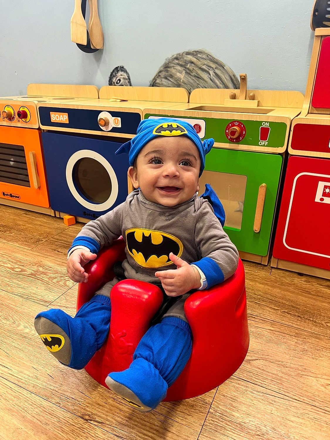 Danny is registered to the contest to win money with this photo: baby, batman_costume, child, smiling, red_baby_seat, wooden_floor, toy_kitchen, colorful, playroom, happy, infant, cute, indoors, costume, headwear, footwear, seat, person, play, young_child