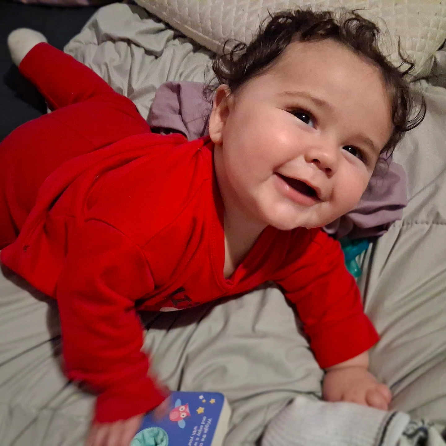 Johnathan joined the competition — help win amazing prizes! baby, bed, blanket, book, child, cozy, curly_hair, cute, face, hand, happy, head, indoor, infant, lying_down, pajamas, person, red_clothing, smiling, toy