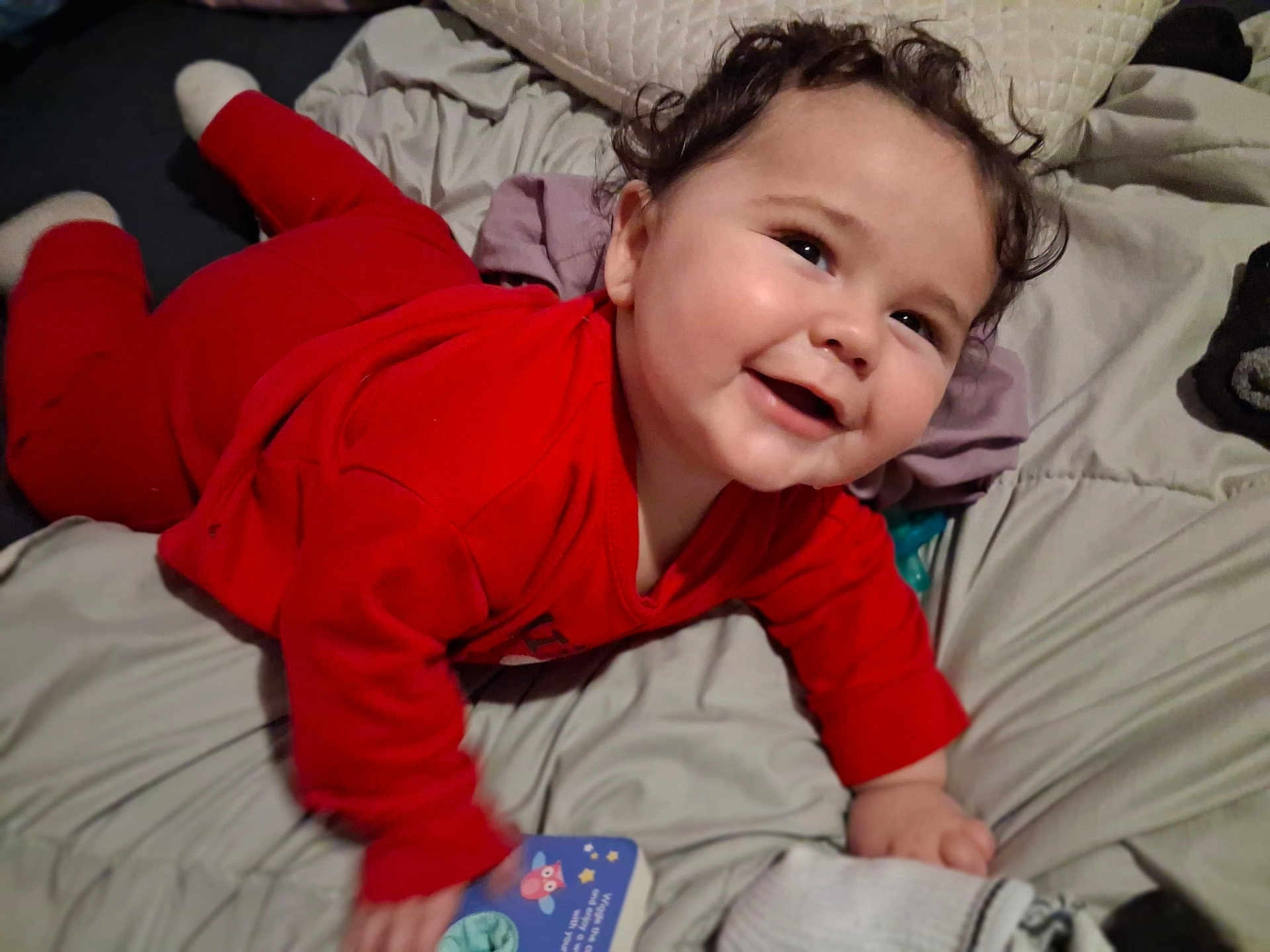 Johnathan joined the competition — help win amazing prizes! baby, child, smiling, red_clothing, bed, blanket, toy, book, happy, indoor, cute, face, hand, head, person, infant, cozy, lying_down, curly_hair, pajamas