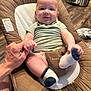 Liam is registered to the contest to win money with this photo: baby, child, infant, person, hand, baby_seat, sock, floor, wood_floor, adult_hand, smiling, green_shirt, indoor, fabric, cushion, home, holding_hand, footwear, cute, comfort
