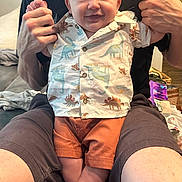 Liam is registered to the contest to win money with this photo: baby, child, smiling, dinosaur_shirt, orange_shorts, adult, holding, indoor, happy, legs, feet, shorts, shirt, person, face, portrait, casual_clothing, home, couch, human