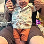 baby, child, smiling, dinosaur_shirt, orange_shorts, adult, holding, indoor, happy, legs, feet, shorts, shirt, person, face, portrait, casual_clothing, home, couch, human