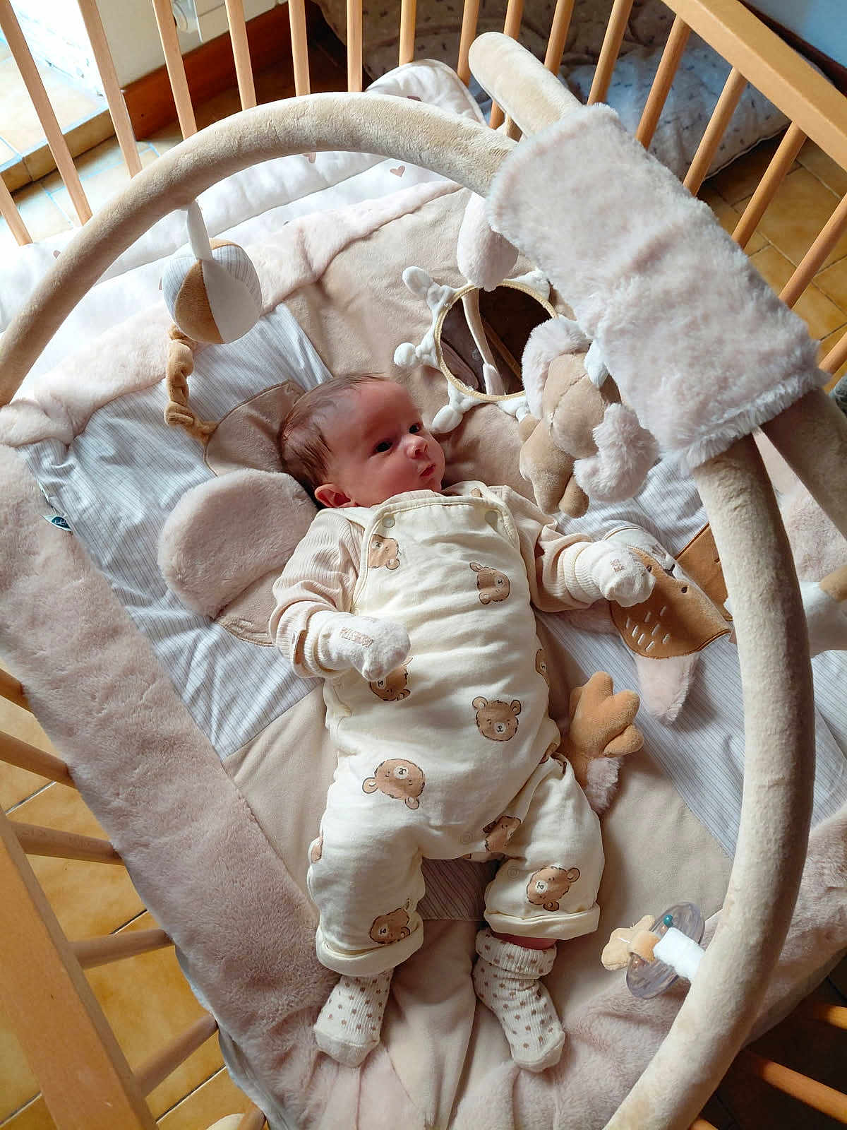 Sohan a rejoint le concours — aidez-le/la à gagner de superbes lots ! baby, crib, plush_toys, pacifier, baby_clothes, mittens, soft_toys, infant, sleeping_area, baby_bedding, wooden_crib, patterned_clothing, cute, indoor, child, floor_tiles, neutral_colors, lying_down, toddler, baby_play_gym