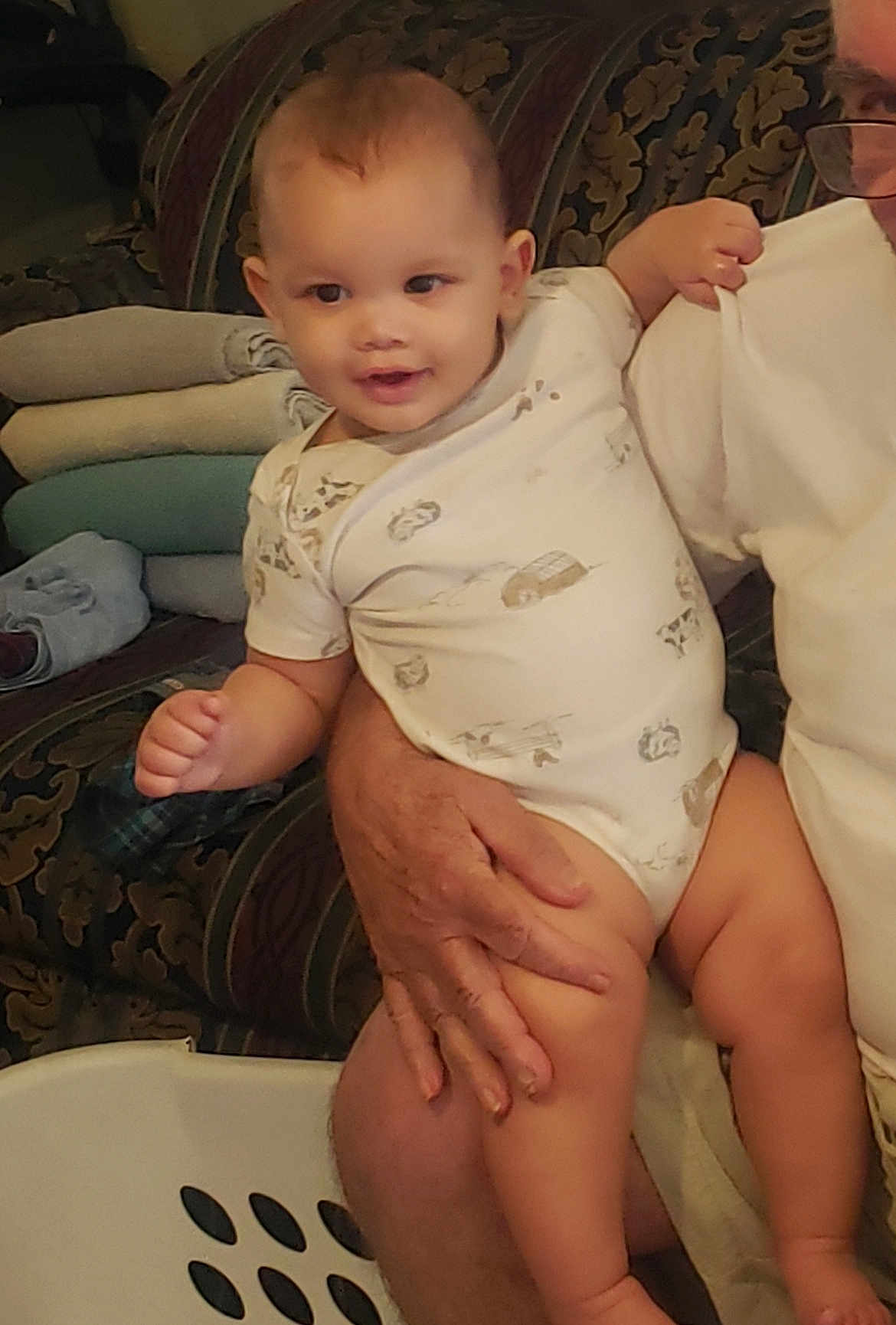 Karson joined the competition — help win amazing prizes! baby, infant, onesie, smile, person, hand, couch, towel, folded_towel, patterned_fabric, indoor, glasses, adult, holding, legs, face, head, happy, skin, home