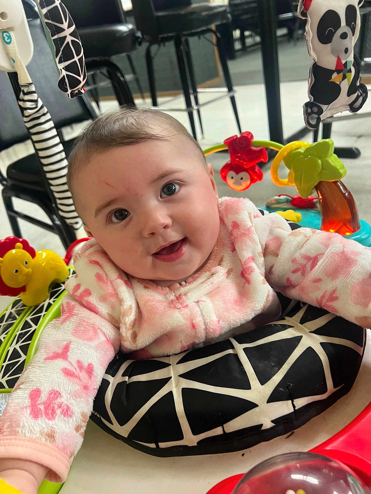 Tinley is registered to the contest to win money with this photo: baby, infant, smiling, eyes, face, pink_clothing, onesie, play_center, toy, lion_toy, panda_toy, activity_center, seat_cushion, indoor, floor, highchair_like, cheek, hair, hand, cute