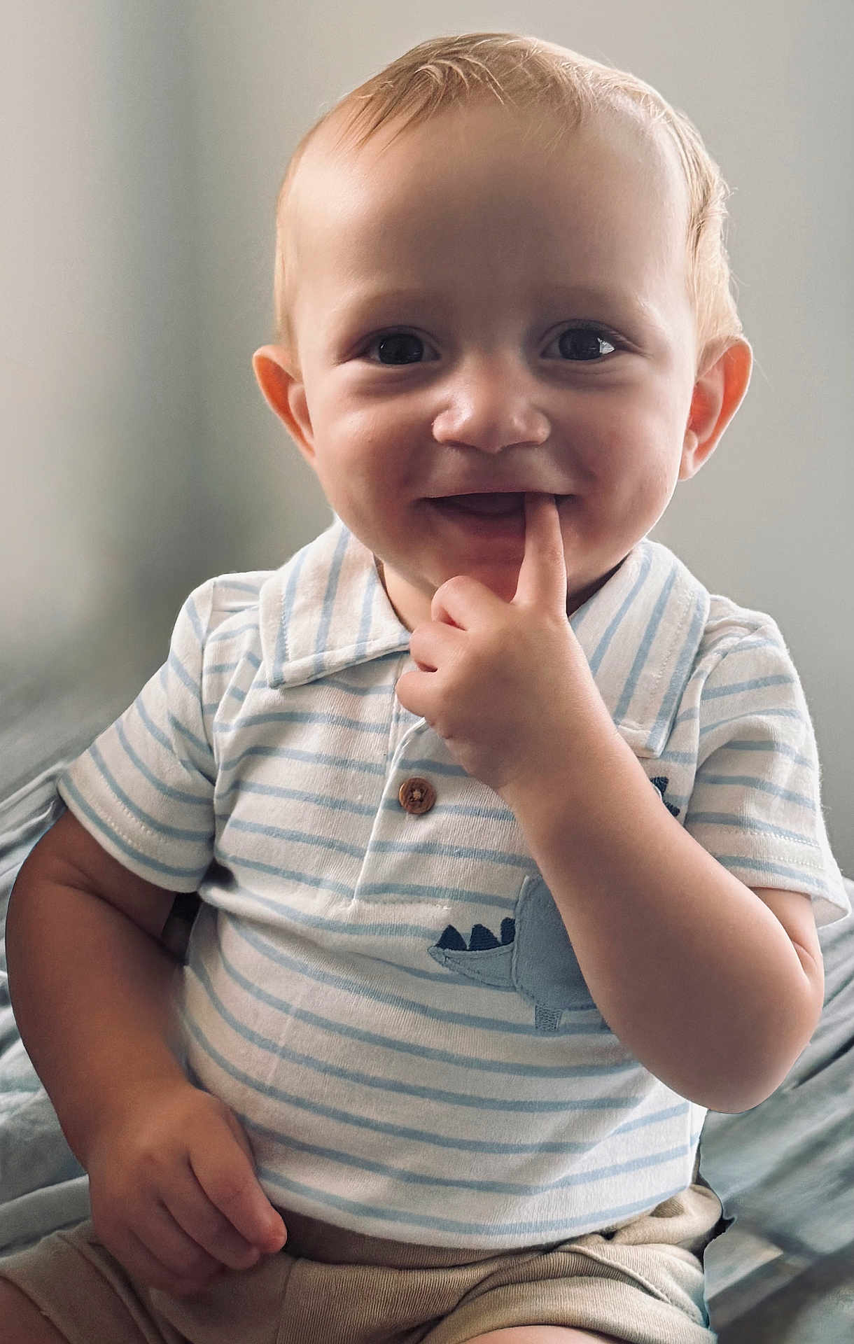 Cash is registered to the contest to win money with this photo: toddler, child, baby, smile, face, hand, finger, striped_shirt, dinosaur_patch, shorts, curly_hair, indoors, natural_light, portrait, cute, playful, sitting, happy, young_child, casual_clothing