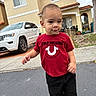 black_shorts, blue_shoes, car, child, clothing, curious, daylight, house, outdoor, person, portrait, red_shirt, residential_area, sidewalk, street, suv, toddler, tree, urban, walking