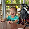 casual, chair, child, daylight, expression, face, glass, greenery, hands, indoor, napkin, person, porch, smiling, stroller, table, toddler, turquoise_shirt, water, wooden_table