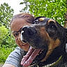dog, person, glasses, outdoor, greenery, trees, smile, tongue_out, happy, selfie, collar, tattoo, nature, face, portrait, canine, animal, closeup, friendship, daylight