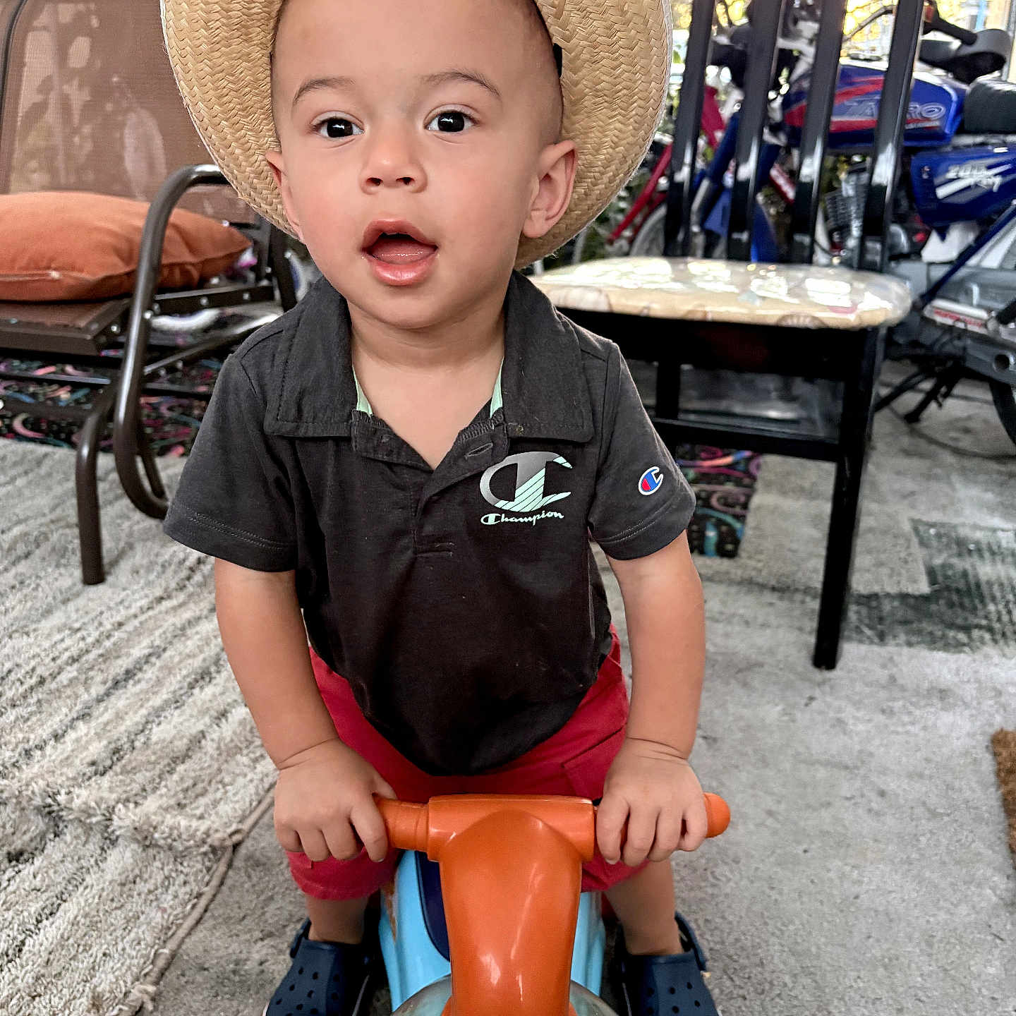 Izael joined the competition — help win amazing prizes! toddler, child, hat, straw_hat, toy_vehicle, indoor, curious, expression, black_shirt, champion_logo, red_shorts, blue_shoes, carpet, chair, plant, floor, face, hands, play, home