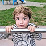 child, boy, striped_shirt, viking_graphic, metal_bar, playground, grass, park, curly_hair, wide_eyes, hands, outdoor, bench, casual_clothing, smiling, cute, person, portrait, daylight, play