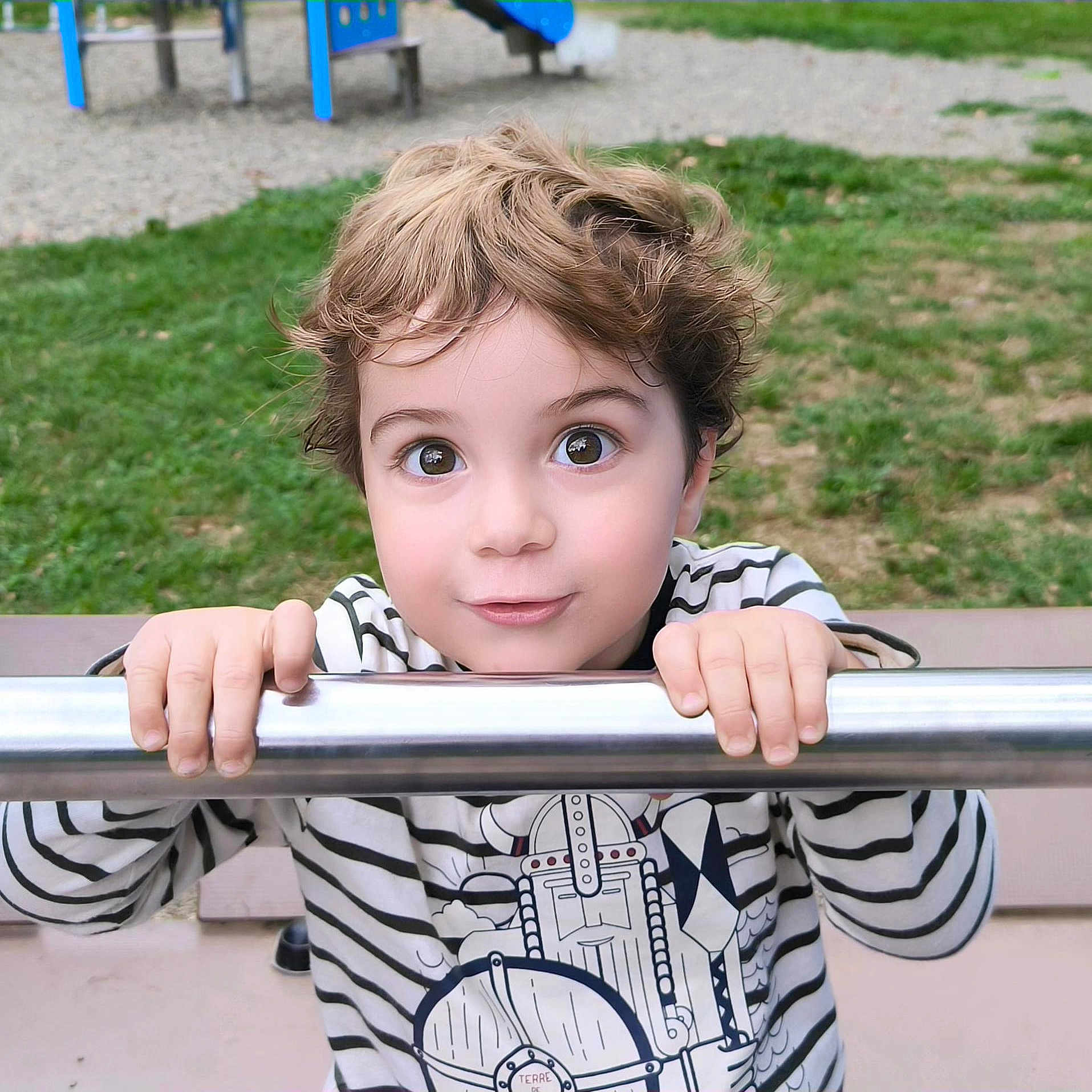 Gabriel participe au concours pour gagner de l'argent avec cette photo : bench, boy, casual_clothing, child, curly_hair, cute, daylight, grass, hands, metal_bar, outdoor, park, person, play, playground, portrait, smiling, striped_shirt, viking_graphic, wide_eyes