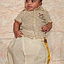 baby, child, traditional_clothing, face, portrait, infant, lying_down, curious_expression, patterned_background, cute, young_child, indoors, textile, gold_trim, short_sleeves, buttoned_shirt, small_hands, head, soft_lighting, cultural